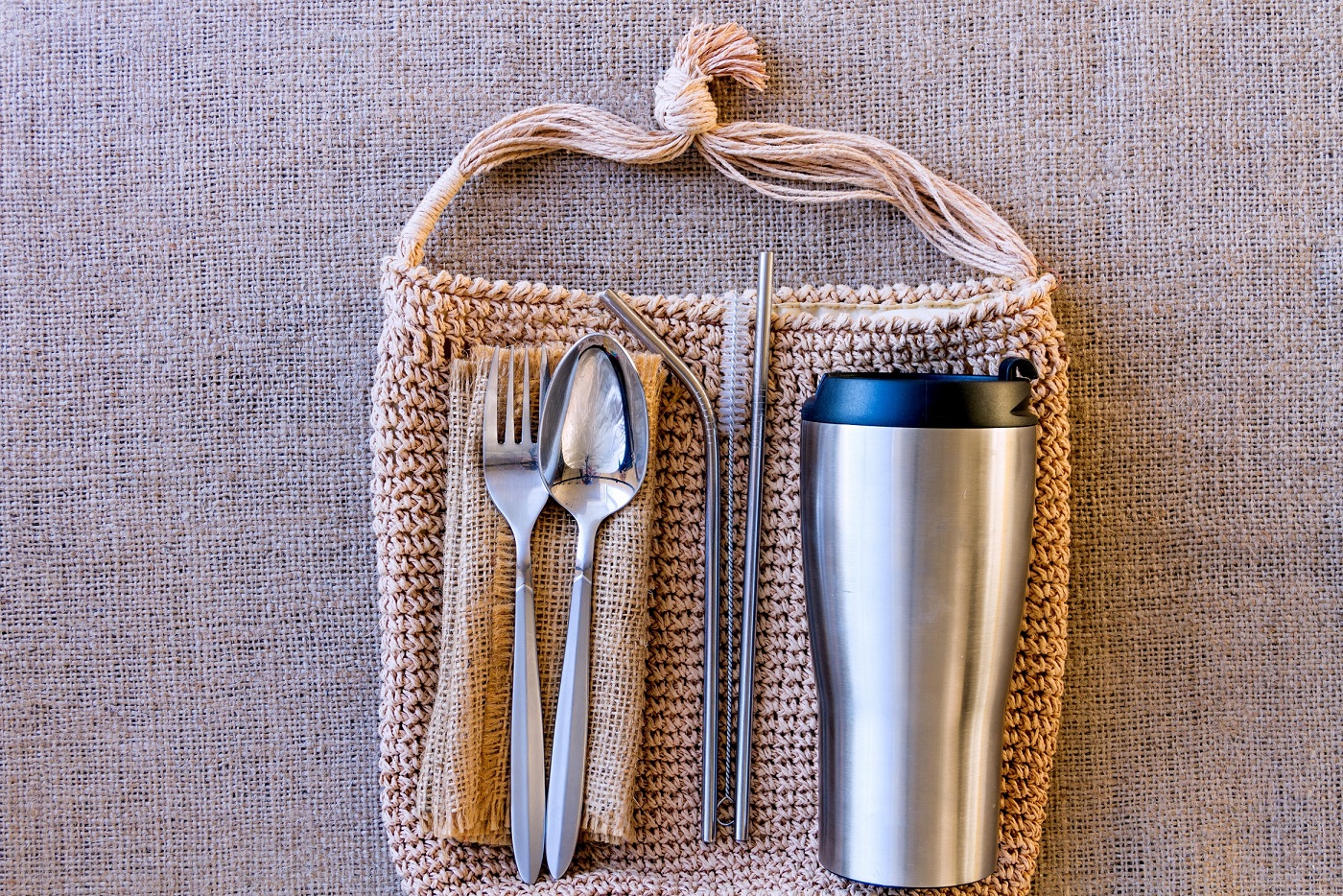 Bag with picnic set on background of burlap. Alternative to disposable tableware. Thermo cup, metal straw with brush, spoon, fork and reusable napkin. Zero waste and plastic free concept.